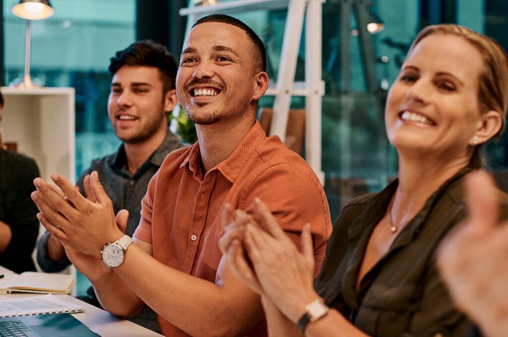 Drei Personen sitzen im Büro und klatschen lächelnd.